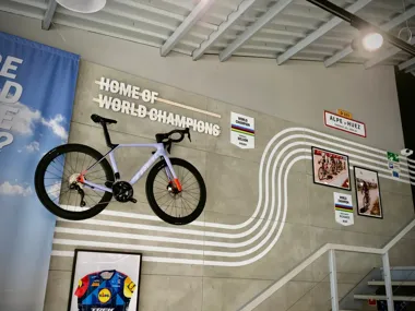 Rower szosowy wiszący na ścianie z napisem „Home of World Champions” i pamiątkowymi tablicami.