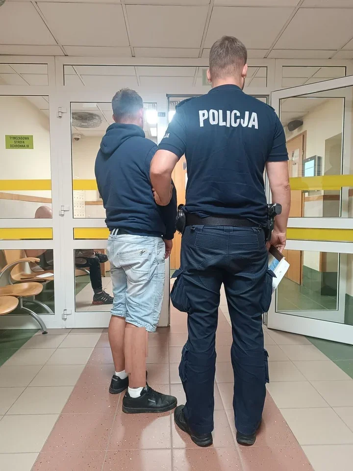 Funkcjonariusz policji w mundurze prowadzi mężczyznę w dresie i szortach przez korytarz budynku z oznaczoną strefą ochronną.