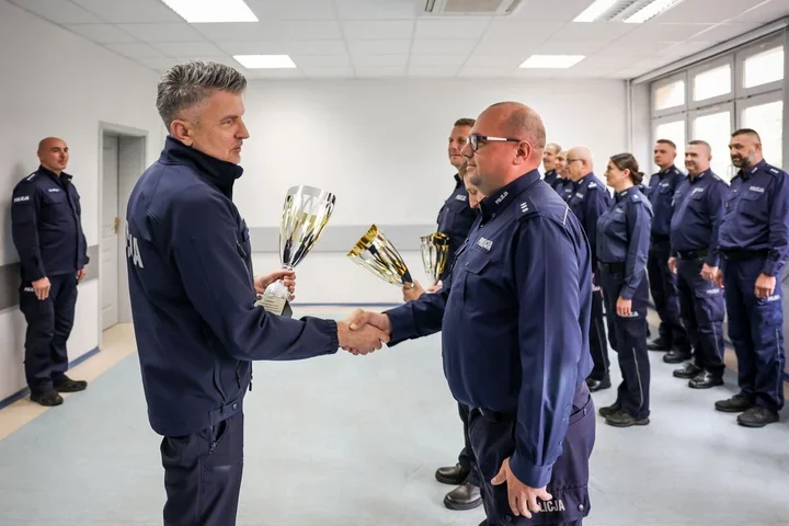 Funkcjonariusze policji w mundurach stoją w sali, a jeden z nich wręcza kolejnemu puchar i podaje mu dłoń.