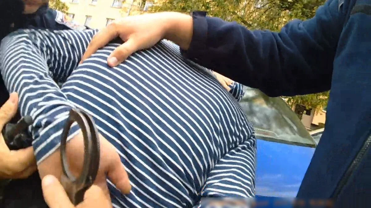 Funkcjonariusz trzyma rękę na plecach mężczyzny w pasiastej koszuli, którego nadgarstki są skrępowane kajdankami, na zewnątrz przy niebieskim samochodzie.