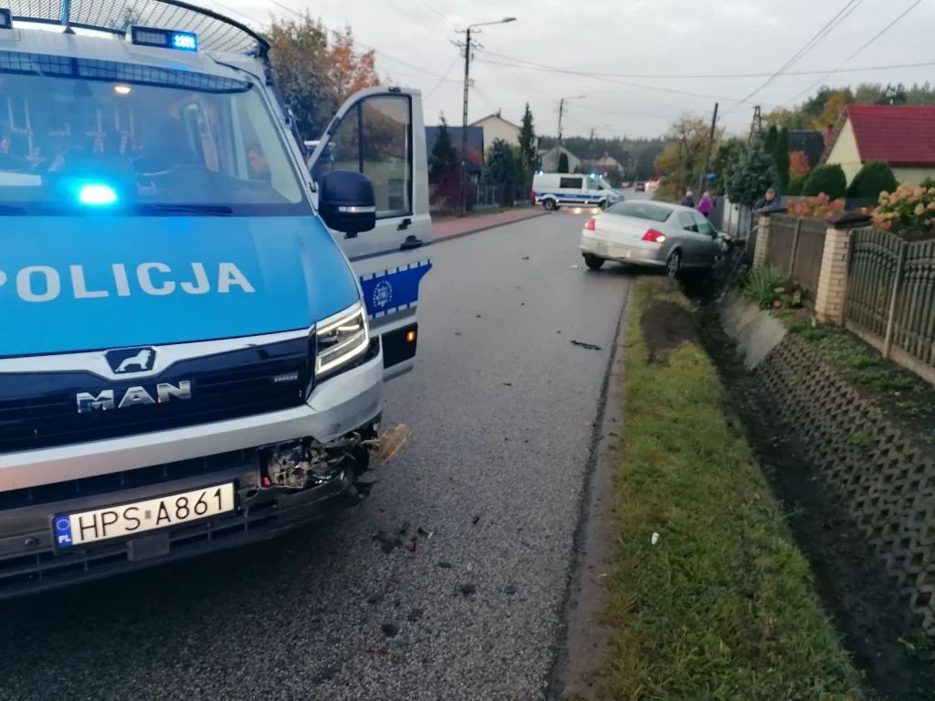 Policyjny MAN z uszkodzonym zderzakiem stoi na drodze wsi obok srebrnego samochodu osobowego, który zjechał do rowu; w tle widać kolejny radiowóz.