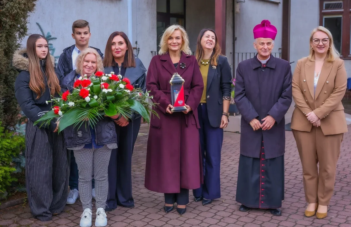 Grupa osób, w tym duchowny w purpurowym kapeluszu, pozuje na brukowanym podwórzu przed budynkiem; jedna z kobiet trzyma wieniec z czerwono-białych kwiatów, a dr