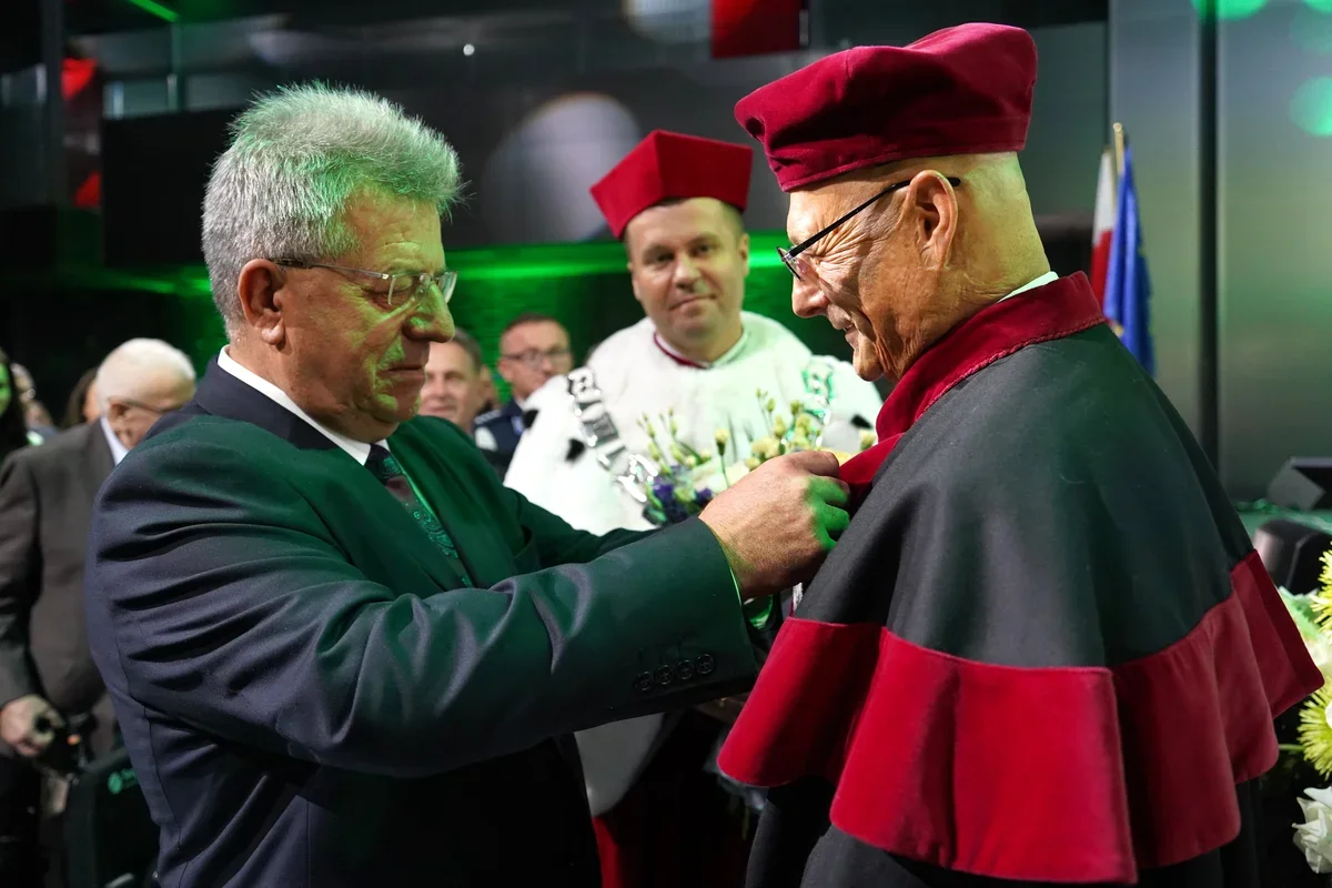 Mężczyzna w garniturze przypina odznaczenie starszemu mężczyźnie w akademickim stroju z bordowym biretem podczas uroczystości.