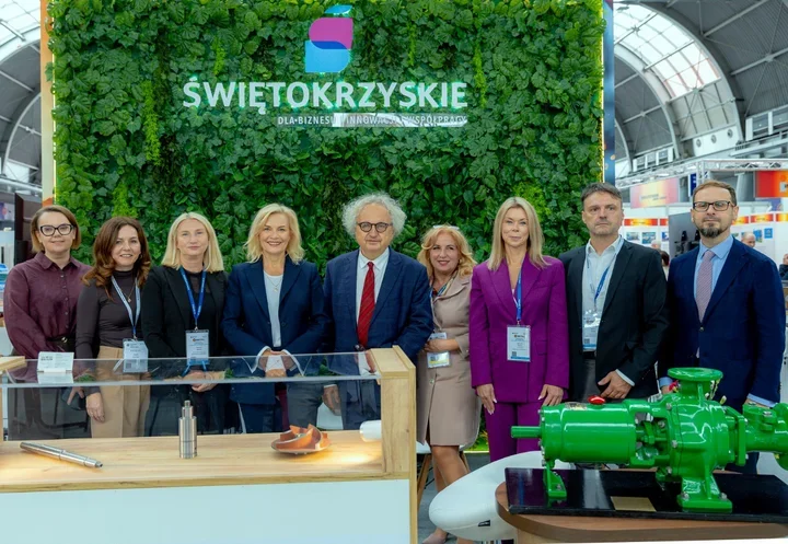 Grupa osób pozuje do zdjęcia przed zieloną ścianą z napisem „Świętokrzyskie” na stoisku targowym z eksponatami przemysłowymi.