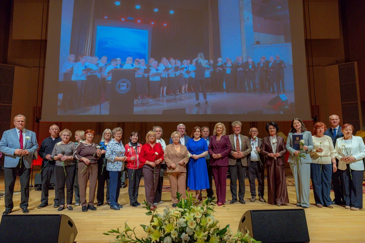 Grupa osób stoi na scenie w auli koncertowej, trzymając kwiaty, z dużym ekranem wyświetlającym zdjęcie grupowe w tle.