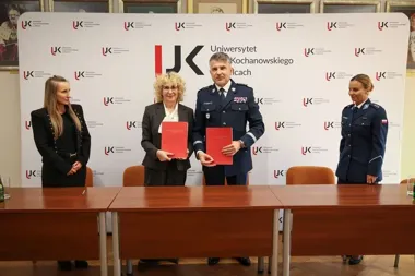 Trzy osoby stojące przy stole na tle ścianki Uniwersytetu Kochanowskiego w Kielcach podczas podpisania porozumienia.