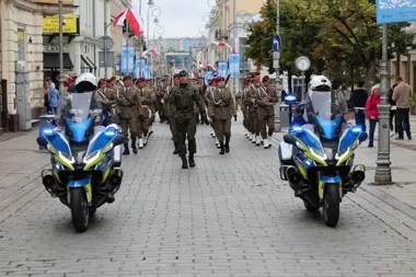 Kolumna żołnierzy w mundurach paradnych maszeruje brukowaną ulicą miasta, a przed nią jadą dwaj policjanci na motocyklach oznakowanych "POLICJA".
