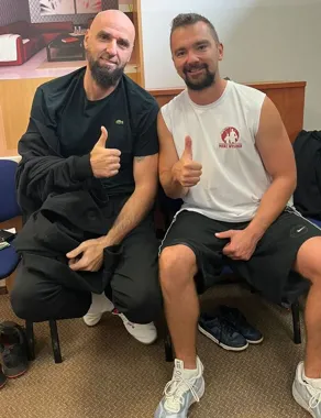 Dwóch mężczyzn siedzi na krzesłach w pomieszczeniu i pokazuje kciuki w górę.