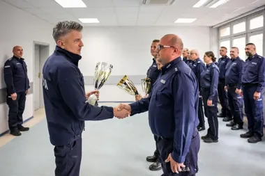 Funkcjonariusze policji w mundurach stoją w sali, a jeden z nich wręcza kolejnemu puchar i podaje mu dłoń.