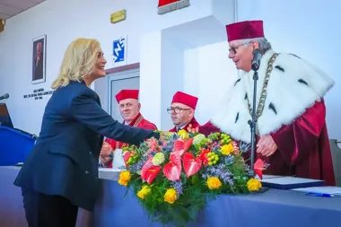 Kobieta w granatowym kostiumie wita się z mężczyzną w bordowej togi i czapce podczas uroczystej akademickiej ceremonii w auli.