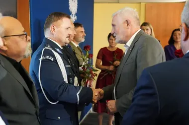 Funkcjonariusz policji w galowym mundurze wita się uściskiem dłoni z mężczyzną w szarym garniturze podczas oficjalnej uroczystości w pomieszczeniu.