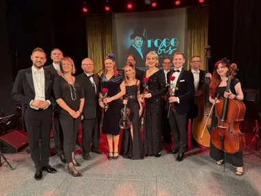 Grupa artystów i muzyków w strojach wieczorowych pozuje na scenie po koncercie, w tle ekran z napisem "FOGG na bis".