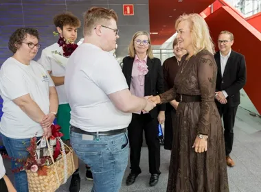 Kobieta w brązowej sukience wita się uściskiem dłoni z młodym mężczyzną w białej koszulce, w otoczeniu kilku osób w nowoczesnym wnętrzu z czerwoną klatką schodo