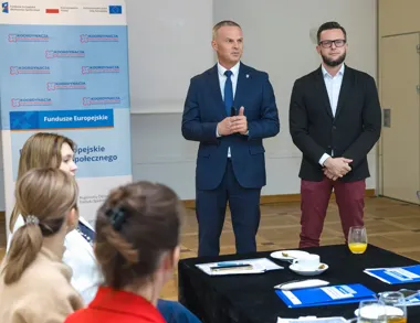 Dwóch mężczyzn w sali konferencyjnej przemawia do grupy siedzących osób przy stole z dokumentami i kubkami, w tle baner z napisami o funduszach europejskich i k