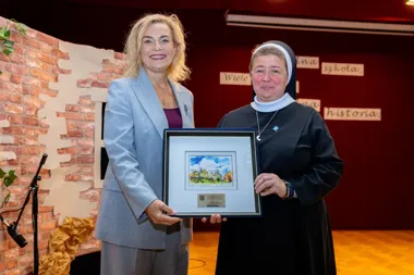 Dwie kobiety, w tym jedna w habicie zakonniczym, stoją na scenie i wspólnie trzymają oprawiony obrazek z widokiem miasta.