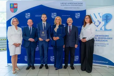 Grupa sześciu osób pozuje do zdjęcia podczas uroczystości w Urzędzie Marszałkowskim Województwa Świętokrzyskiego, mężczyzna w centrum trzyma dokument z napisem