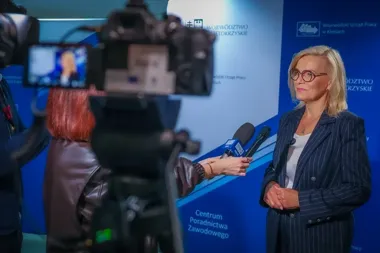 Kobieta w prążkowanym garniturze udziela wywiadu dziennikarce z mikrofonem TVP3 przed kamerą w siedzibie Wojewódzkiego Urzędu Pracy w Kielcach.