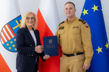 Kobieta w prążkowanym garniturze wręcza dokument oznaczony napisem „Marszałek Województwa Świętokrzyskiego” funkcjonariuszowi Państwowej Straży Pożarnej, na tle
