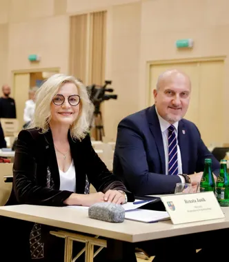 Kobieta i mężczyzna siedzą przy stole w sali konferencyjnej, przed nimi leżą dokumenty i tabliczka z nazwiskiem Renata Janik.
