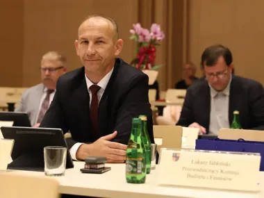 Mężczyzna w garniturze siedzi przy stole konferencyjnym z tabletem, butelkami wody i tabliczką z nazwiskiem, w tle inni uczestnicy spotkania.