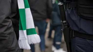 Fragment sylwetki kibica w zielono-białym szali obok policjanta z pałką w kaburze.
