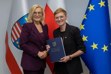 Dwie kobiety w garniturach stoją przed flagami województwa świętokrzyskiego, Polski i UE, jedna wręcza drugiej dokument z napisem Zarząd Województwa Świętokrzys