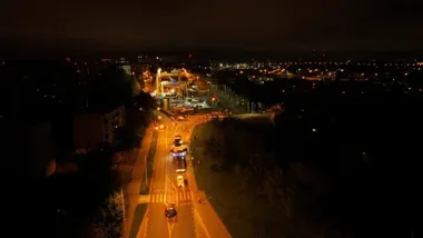 Nocne ujęcie z lotu ptaka na oświetloną ulicę z pojazdami, w tym ciężarówką przewożącą duży ładunek, oraz miasto w tle.