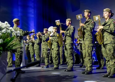 Wojskowa orkiestra dęta w mundurach polowych występuje na scenie.
