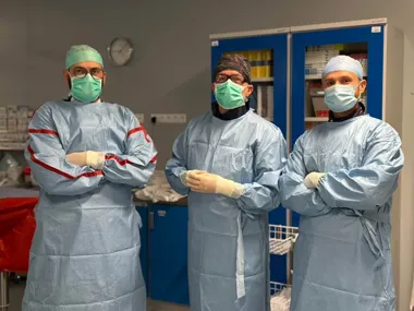 Trzech członków zespołu medycznego w fartuchach chirurgicznych, maskach i rękawiczkach stoi w pomieszczeniu szpitalnym przy szafkach z medycznymi zaopatrzeniem.