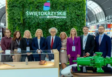 Grupa osób pozuje do zdjęcia przed zieloną ścianą z napisem „Świętokrzyskie” na stoisku targowym z eksponatami przemysłowymi.