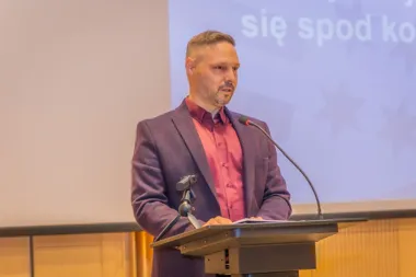 Mężczyzna w fioletowej marynarce i czerwonej koszuli przemawia z mównicy w sali konferencyjnej, za nim ekran z projekcją.